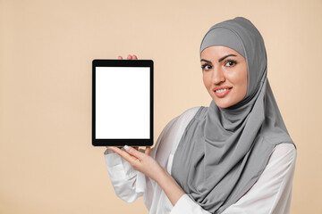 Young muslim Middle Eastern islamic arabian woman showing digital tablet`s screen isolated over beige background. Smart student in grey hijab using tablet for online studing, webinars, chatting
