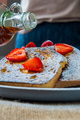 Woman pouring maple syrup on a french toast plate with fresh strawberries, raspberries and powdered sugar. Tasty morning breakfast or brunch with gluten free bread. Easy making and healthy dessert.