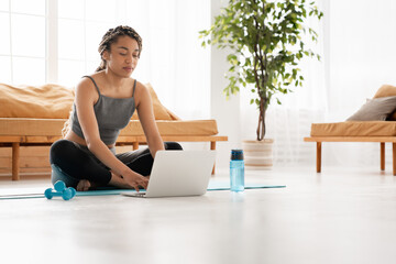 Young slim african girl sitting in lotus position at home using laptop. Cheerful African woman...