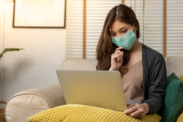 Cute young asian girl working on a lap top from the comfort of her home. She is wearing sweater sitting on a couch. She is wearing face mask.
