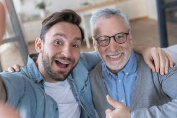 Cheerful caucasian adult son and old senior father taking making selfie at home, spending time...