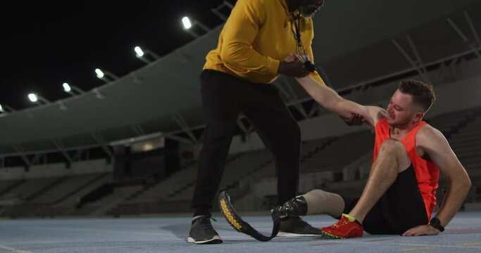 Diverse male coach helping tired disabled male athlete with running blade to stand up