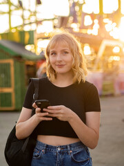 Mujer joven usando su móvil durante la puesta de sol en una feria o parque de atracciones