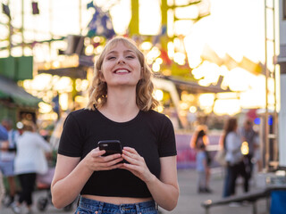 Mujer joven usando su móvil durante la puesta de sol en una feria o parque de atracciones