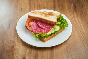 Delicious snack - toast sandwich with salami, cucumber, tomatoes and sauce in a white plate on a wooden background