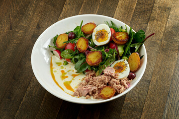 Classic nicoise salad with boiled egg, potatoes, tuna, olives and white sauce, served in a white bowl on a wooden plate. A delicious and healthy seafood salad