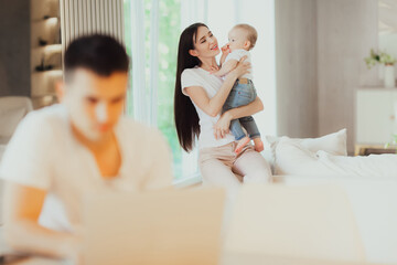 Fototapeta premium Young mother looks after her baby while her husband works on laptop