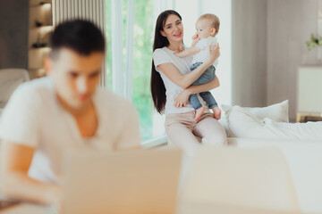 Fototapeta premium Young mother looks after her baby while her husband works on laptop