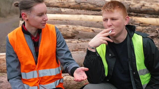 Sawmill workers talking. Smoking cigarettes at leisure. Taking a break from work, recuperating. Workers pause logging work. They sit on logs, smoke a cigarette. 