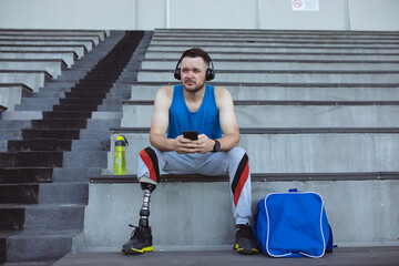 Caucasian male athlete with prosthetic leg using smartphone sitting on the seats in the stadium