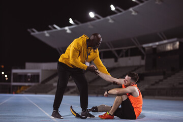 African american male coach helping caucasian male athlete with prosthetic leg to get up on track
