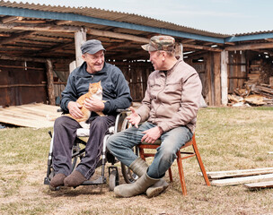 A happy senior man in work clothes in a wheelchair in the yard of his house, talking to his friend.