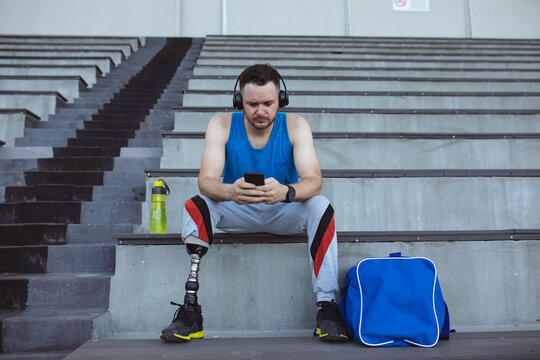 Caucasian male athlete with prosthetic leg using smartphone sitting on the seats in the stadium