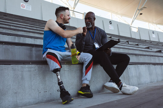 African american male coach and caucasian male athlete with prosthetic leg fist bumping in stadium - Powered by Adobe