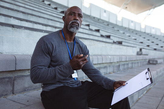 African american male coach with stopwatch measuring time while sitting on the seats in the stadium