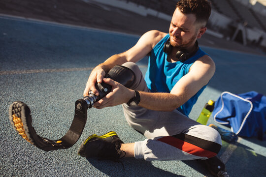 Caucasian male athlete fixing his prosthetic leg while sitting on running track in the stadium