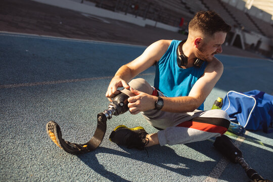 Caucasian male athlete fixing his prosthetic leg while sitting on running track in the stadium