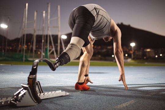 Caucasian male athlete with prosthetic leg in starting position for running on the track at night
