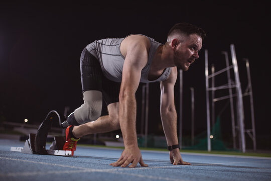Caucasian male athlete with prosthetic leg in starting position for running on the track at night