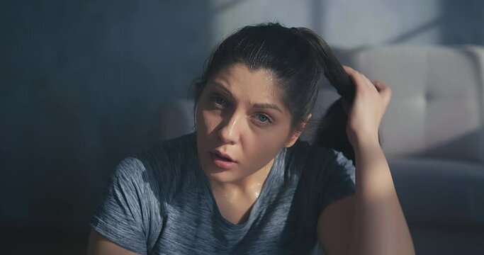 Tired sportswoman with sweaty face wearing grey t-shirt strokes hair resting after intensive training in room closeup slow motion