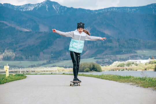 Cute girl skateboarding on the longboard on the asphalt road with a mountain landscape background. She gracefully balancing when slow riding. Happy outdoor childhood concept image.
