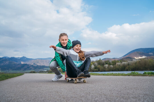 Laughing kids have fun using a skateboard. Brother pushing his sister sitting on a skateboard and making wings with arms. Happy outdoor childhood without an electronic devices concept image.