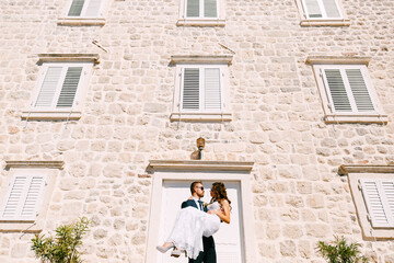 Fototapeta na wymiar Groom holding bride in his arms against the stone wall of the building on a bright sunny day.
