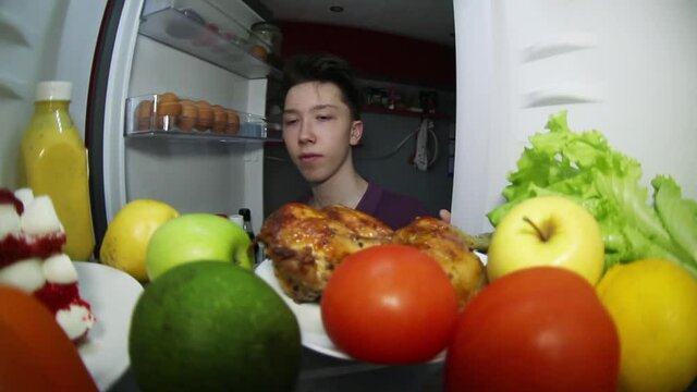 The teenager looks in the refrigerator for food. Makes a choice: smoked chicken or fruit with vegetables, or cake on a plate.