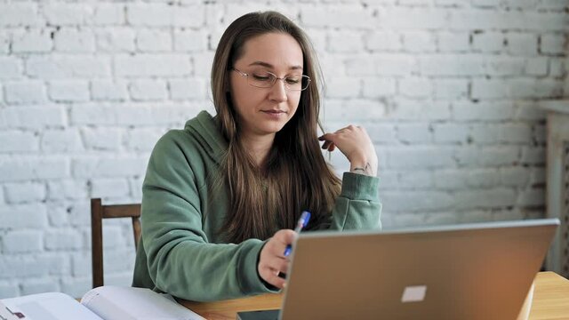 Smiling female student in glasses elearning study with online tutor teacher talk conference calling do video chat learn language make notes sit at home office desk look at laptop computer