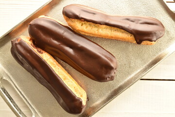 Three chocolate eclairs on a metal tray, close-up, on a wooden table, top view.