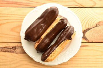 Three chocolate eclairs on a white ceramic plate, close-up, on a wooden table, top view.