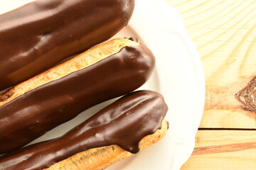 Three chocolate eclairs on a white ceramic plate, close-up, on a wooden table, top view.