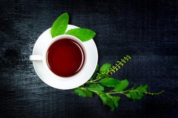 Tulsi or holy basil tea in white cup with fresh tulsi branch and leaf on black wooden background, top view, darktone. Ayurvedic medicine in India.