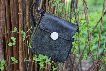 old black leather bag hanging on brown branches of a bush with green leaves outside in the garden