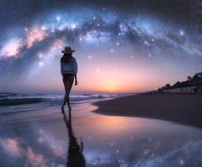Arched Milky Way and young woman in hat on sandy beach against starry sky reflected in water at...