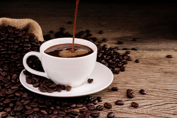 A white coffee cup, complete with a large number of black coffee and roasted coffee beans, is laid around on a wooden table in a warm atmosphere. On a dark background with a copy area
