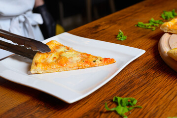 Close up of chef placing hot cheesy slice of pizza slice on square white plate using serving tongs.