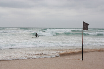 A surfer enters a stormy sea against the backdrop of a black flag that prohibits swimming.