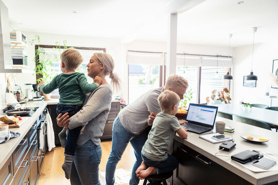 Mother carrying sin while father using laptop with elder sibling in kitchen