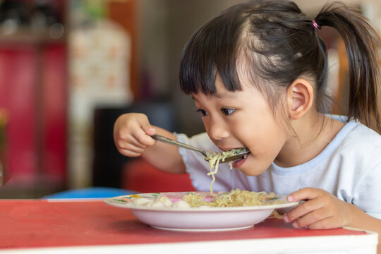 Portrait​ image​ of​ 3-4 years​ old​ of​ baby.​ Happy​ Asian​ child​ girl​ enjoy​ eating​ some​ noodles by​ herself.​ Healthy​ Food​ and​ kid​ concept.