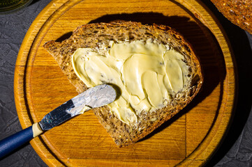 Slice of rustic natural yeast-free bread with flax, poppy seeds, sesame seeds, millet, pumpkin and sunflower seeds, spread with butter, on a wooden board, black background, hard light