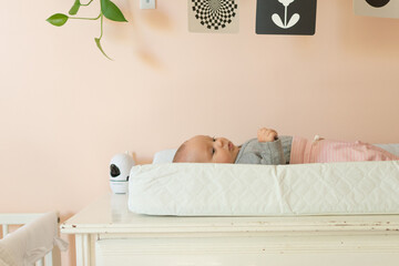Young baby laying on changing pad in a peach nursery; black and white mobile and baby monitor nearby