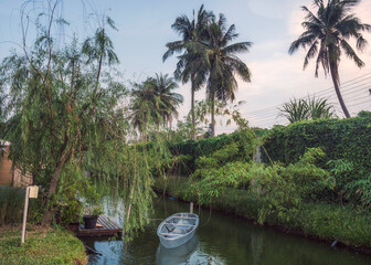 Rural garden with transparent boat by wooden pier on the canal at evening