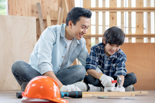Asian father look carefully son use hammer to hopnailed in their workplace of carpentering with happy emotion. Asian family concept to stay at home and enjoy good relationship hobby together.