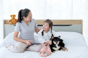 Asian mother and little girl look at face each other and smile with happiness and stay on bed with their dog.