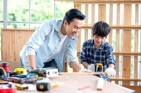 Asian father look carefully son use saw with timber in their workplace of carpentering with happy emotion. Asian family concept to stay at home and enjoy good relationship hobby together.