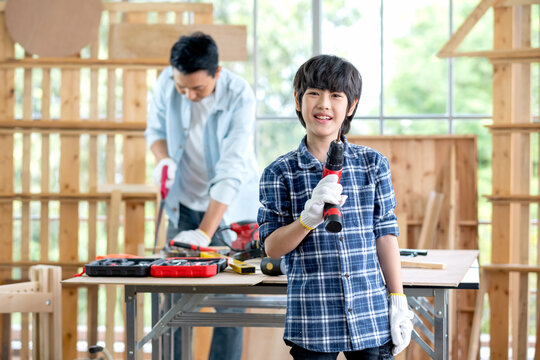 Asian boy hold electric drill and smile with look at camera. His father stay on the background and work as carpentering at home.