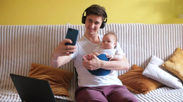 Young freelance father makes a photo on the phone. He is holding a toddler in his arms. Spending time with your child. Headphones are on the head.