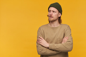 Portrait of handsome, adult male with blond hair and beard. Wearing green beanie and beige sweater. Keeps arms crossed. Watching to the left at copy space, isolate over yellow background