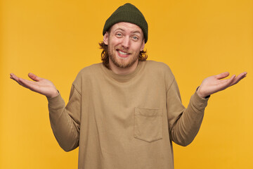 Portrait of doubtful, confused male with blond hair and beard. Wearing green beanie and beige sweater. Shrugs with hands lifted and wry face. Watching at the camera isolated over yellow background